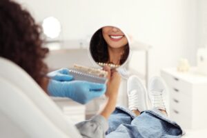 Patient looking in mirror while doctor holding teeth color chart in office, closeup. Dental veneers cosmetic procedure 