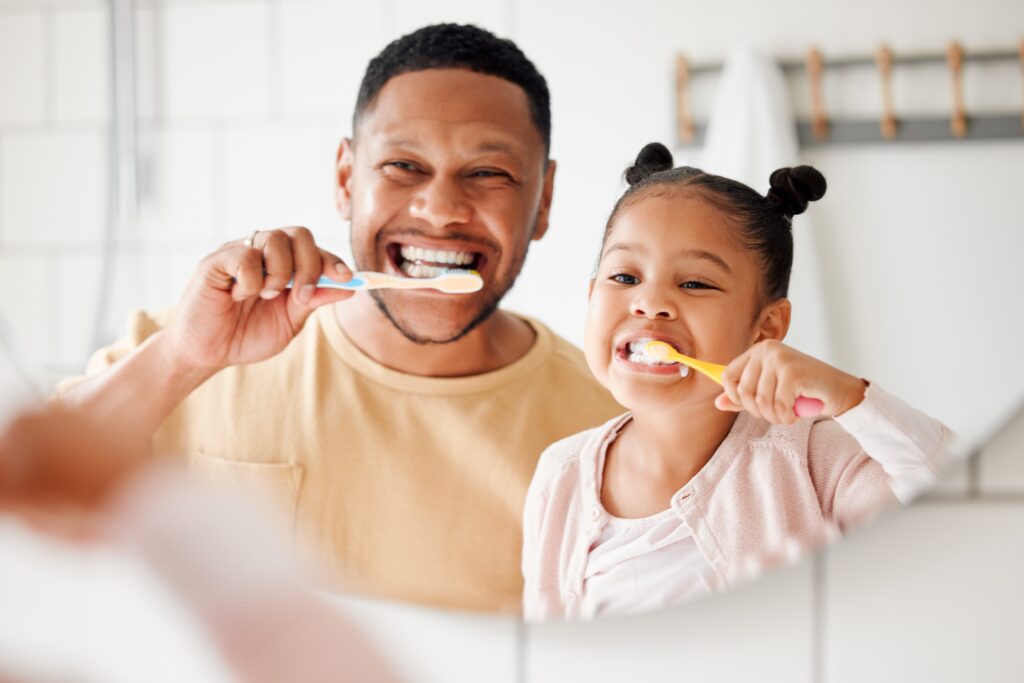 father and daughter brushing their teeth in the mirror, practicing proactive dental care