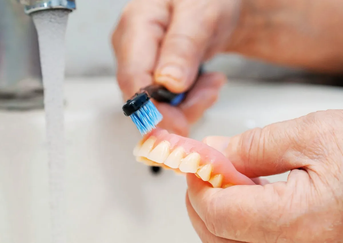 Person brushing removable dentures to help maintain their condition and extend denture lifespan.